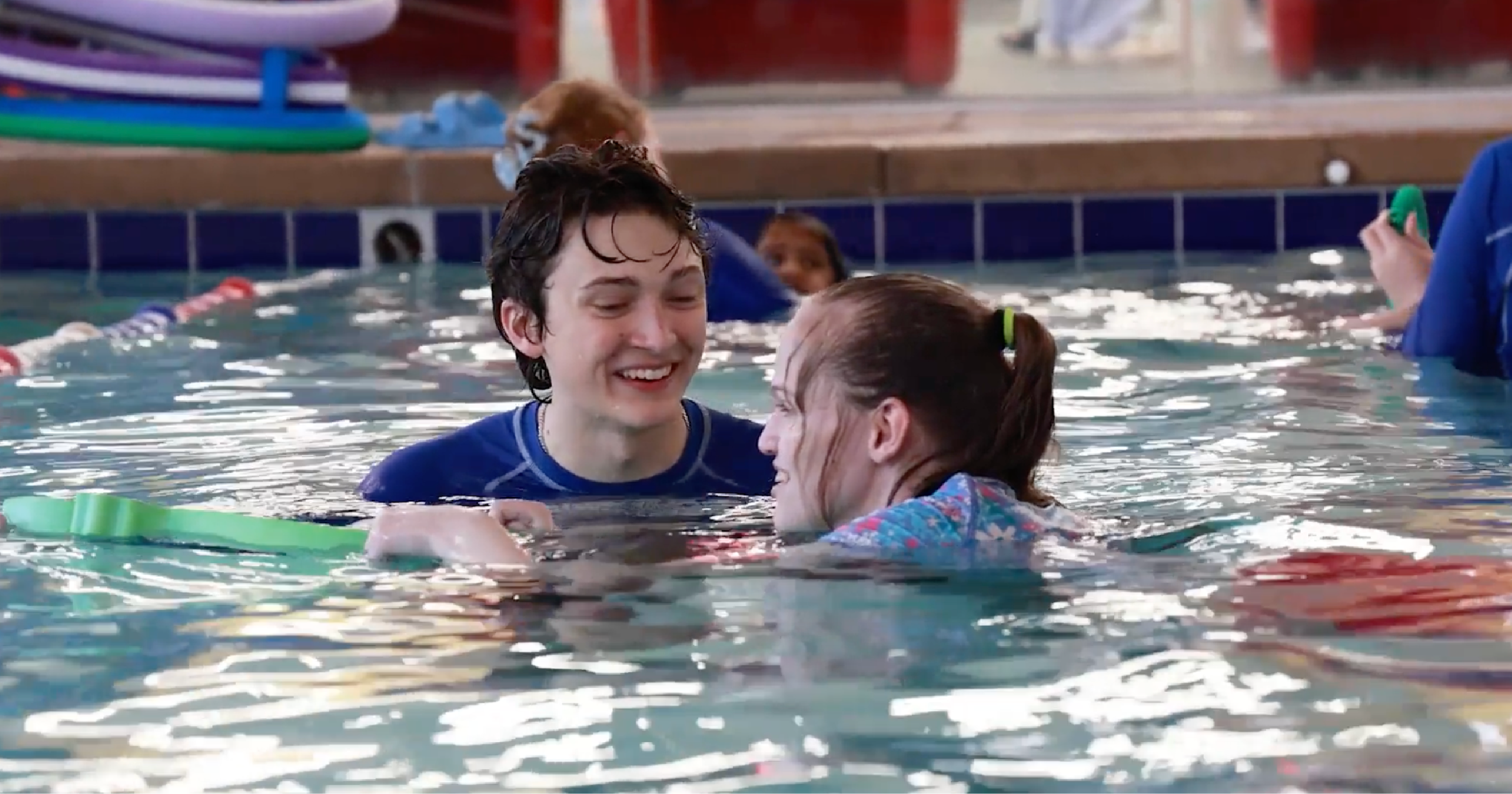 Image of Lucy Cook, a teen with cerebral palsy, using a kickboard in the pool while Cory, her Aqua-Tots instructor, is standing next to her, smiling and guiding her through the adaptive swim lesson.